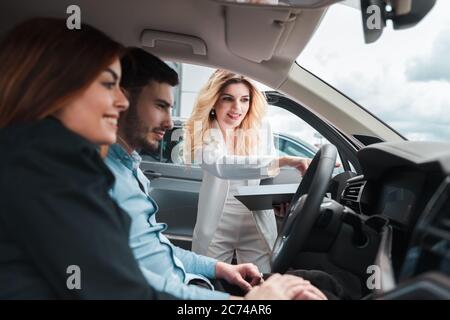 Junges Paar im Auto. Eine Beraterin zeigt einem jungen Paar ein Auto. Junge Familie wählt Auto bei einem Autohaus Stockfoto