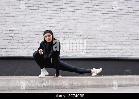 arabischer Sportlerin im Hijab, der sich in der Nähe einer Ziegelmauer erstreckt Stockfoto
