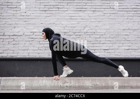 Seitenansicht der arabischen Sportlerin in Hijab, die sich in der Nähe einer Ziegelmauer erstreckt Stockfoto