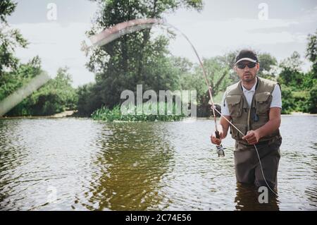 Junge Fischer fischen auf See oder Fluss. Ernst konzentrierter Kerl in Fischer Kleidung stehen im Fluss oder See Wasser und halten Stange. Es wird versucht Stockfoto