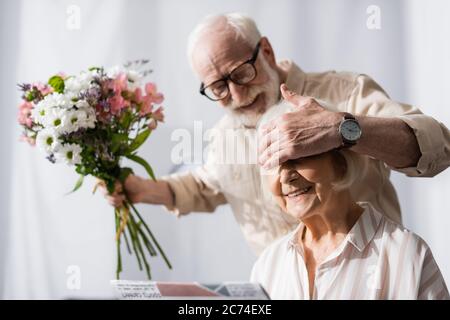 Selektiver Fokus auf lächelnder älterer Mann, der die Augen der Frau bedeckt und Bouquet hält Stockfoto