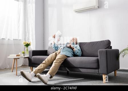 Müde älterer Mann mit Zeitung auf der Couch unter Klimaanlage zu Hause Stockfoto