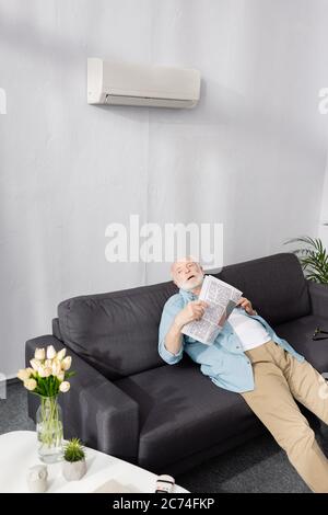 Erschöpfter älterer Mann, der Zeitung unter der Klimaanlage auf der Couch hielt Stockfoto