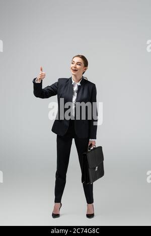 Fröhliche junge Geschäftsfrau mit Lederkoffer zeigt Daumen nach oben isoliert auf grau Stockfoto