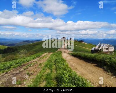 Erdige leere Straße entlang der Spitze der Karpaten hoch unter weißen geschwollenen Wolken. Holzhäuser am Horizont und am Straßenrand. Stockfoto