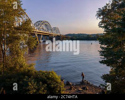 Männchen wirft an einem Sommerabend eine Angelrute am Ufer des Flusses Dnipro in der Nähe der riesigen Eisenbahnbrücke. Urban Hobby und aktiver Lebensstil im Freien. Stockfoto