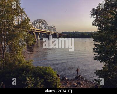 Männchen wirft an einem Sommerabend eine Angelrute am Ufer des Flusses Dnipro in der Nähe der riesigen Eisenbahnbrücke. Urban Hobby und aktiver Lebensstil im Freien. Stockfoto