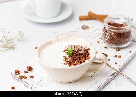 Joghurt mit Schokoladengranola in Tasse, Frühstück mit Tee auf weißem Holzhintergrund, Seitenansicht. Stockfoto