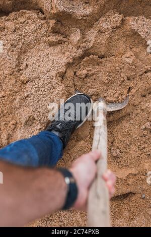 Erste-Person-Ansicht einer Schaufel in den Händen eines Arbeiters - Sammeln von Sand aus einem großen Haufen auf einer Baustelle Stockfoto