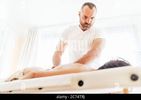 Bärtiger Masseur, der eine therapeutische Rückenmassage für ein Mädchen macht, das auf einer Massagesuht in einem Massagewabor liegt. Stockfoto