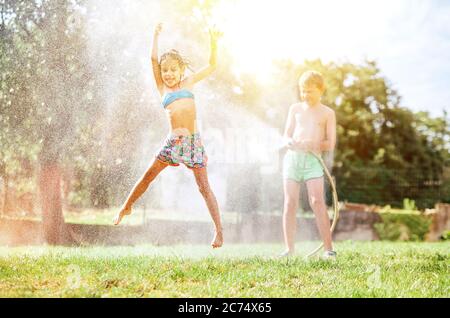 Glückliches kleines Mädchen springt unter Wasser, als Bruder sie aus Gartenschlauch gießt. Heiße Sommertage Aktivität. Stockfoto