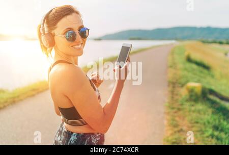 Junge schöne Frau beginnt Joggen und Musik hören mit Smartphone und kabellosen Kopfhörern fröhlich lächelnd in der Kamera Stockfoto