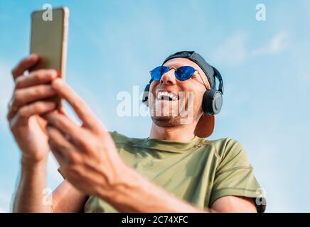 Bärtiger Mann in Baseballmütze, kabellose Kopfhörer und blaue Sonnenbrille fanden etwas Lustiges in Smartphone-Gerät und fröhlich lachen. Stockfoto