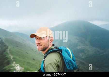 Müde lächelnder Backpacker Mann in Baseball-Cap zu Fuß durch die neblig bewölkten Wetter Bergkette Pfad mit Rucksack. Aktiv Sport Rucksackwandern gesund Stockfoto