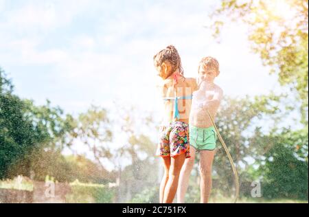 Bruder spuckt seine kleine Schwester Wasser aus Gartenschlauch. Kinder haben Spaß in heißen Sommertag Stockfoto