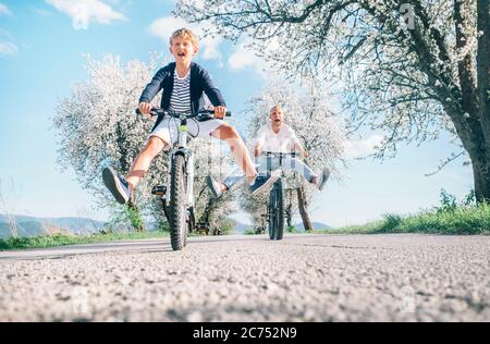 Vater und Sohn haben Spaß, breiten Beinen und Schreien beim Radfahren auf der Landstraße unter Blütenbäumen. Gesundes sportliches Lifestyle-Konzept Stockfoto