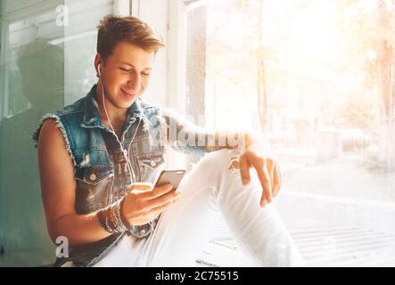 Junger Mann hört Musik mit Kopfhörern und Smartphone auf Fensterbank sitzen Stockfoto