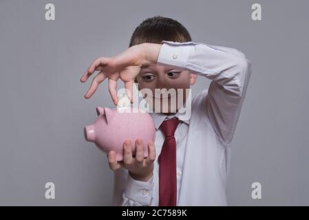 Lächelnder Junge steckt eine Münze in ein rosa Sparschwein. Geschäftskonzept. Heller Hintergrund Stockfoto