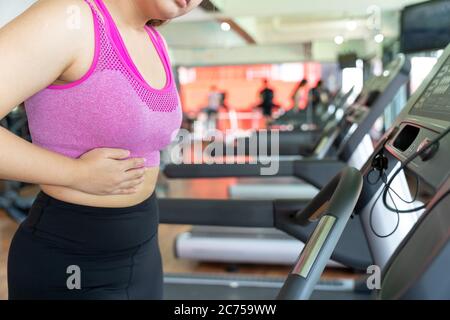 Eine Frau, die sich erschöpft fühlt und an Magenschmerzen und Verletzungen leidet, während sie auf dem Laufband im Fitnessstudio läuft. Sport, Gesundheitswesen und medizinische Co Stockfoto