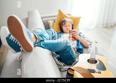 Pre teen Junge mit Gitarre auf gemütlichen Sofa gekleidet Casual Jeans und neue Sneakers Musik hören und chatten mit drahtlosen Kopfhörern verbunden w Stockfoto