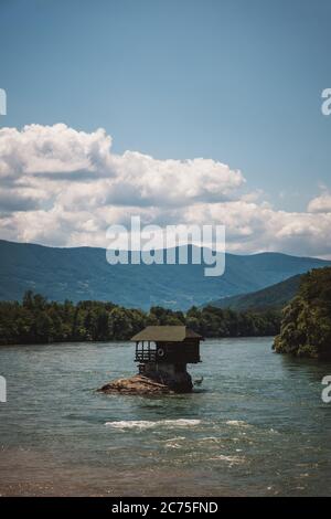 Hölzerne Drina River House steht auf einem Felsen in der Mitte des Flusses. Dahinter liegen Berge und blauer Himmel mit Wolken an einem sonnigen Tag. Vertikales Bild Stockfoto