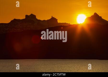 Schöner Sonnenuntergang in felsigen Bergen und Seeufer, rot orange Sonne untergeht hinter Silhouette der Klippe Bergkette, malerische Landschaft Stockfoto