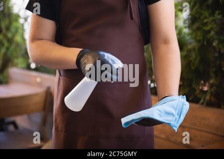 Der Kellner arbeitet in einem Restaurant in einer medizinischen Maske, Handschuhe während Coronavirus Pandemie. Stellt eine neue Norm in Service und Sicherheit dar. Desinfizierung von Tischen und Oberflächen. Betreuung der Kunden. Stockfoto