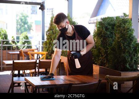 Der Kellner arbeitet in einem Restaurant in einer medizinischen Maske, Handschuhe während Coronavirus Pandemie. Stellt eine neue Norm in Service und Sicherheit dar. Desinfizierung von Tischen und Oberflächen. Betreuung der Kunden. Stockfoto