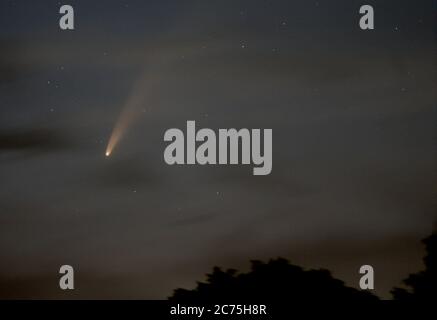 Komet C/2020 F3 NEOWISE leuchtet in der Nacht vom 12. Juli 2020 hell hinter Wolken und trägt einen langen Staubschweif und ein helles Koma nahe dem Nordhorizont. Stockfoto