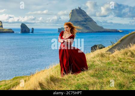 Eine junge Rotschopf-Frau bleibt in altmodischer Kleidung auf einer Klippe nahe der Insel Tindholmur. Atlantikküste, Insel Vagar, Färöer Inseln. Europa Stockfoto