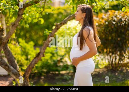 Nachdenkliche schwangere Frau, die im Garten steht und die Hände auf dem Rücken hält Stockfoto