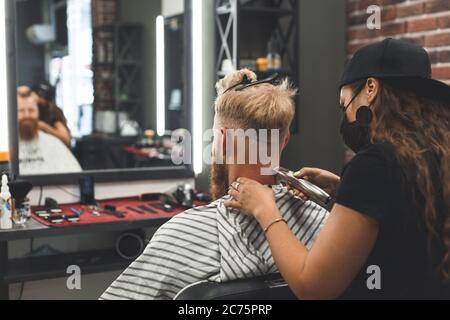 Weibliche Barbier in Maske schneidet einen Mann Haare mit Haarschneider. Frisur während der sozialen Distanzierung Stockfoto