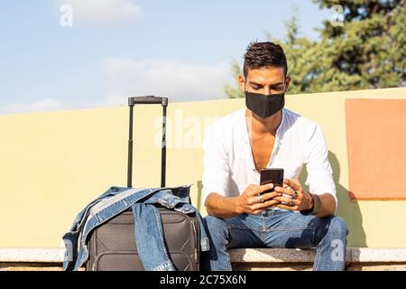 Junge stilvolle kaukasischen Mann Reisende mit einer schwarzen COVID Schutzmaske mit dem Smartphone sitzt auf einer bunten Bank mit der Denim-Jacke auf einem tr Stockfoto