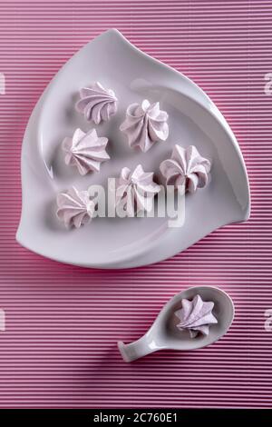 Flach mit zarten rosa Baiser-Plätzchen. Wenige Bagues sind auf weißem Porzellanteller und einer ist in Porzellanlöffel auf rosa gestreiftem Papier backgroun Stockfoto