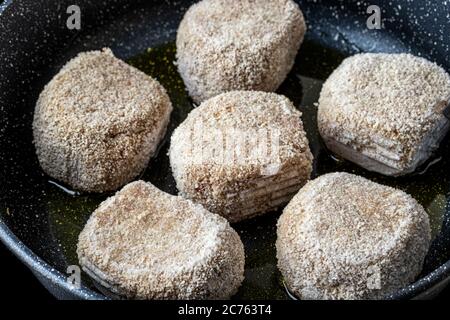 Rohe gefrorene panierte Hähnchenschnitzel, in einer schwarzen Keramikpfanne mit Pflanzenöl erhitzt. Der Prozess des Bratens von Steaks. Traditionelle Küche. Stockfoto