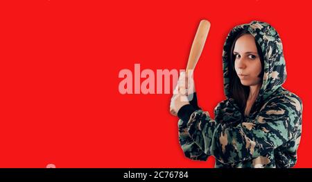 Portrait einer jungen Frau im Camouflage Sweatshirt mit Fledermaus in den Händen. Aggressiver Schläger in der Kapuze mit Baseballschläger auf rotem Hintergrund. Stockfoto