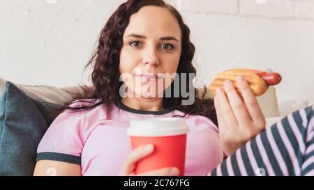 Eine junge Frau im Bett, die einen Hot Dog isst und Kaffee trinkt. Weibchen isst einen saftigen Hot Dog in einem gemütlichen Schlafzimmer. Konzept: Versuchung auf Diät und Fast-Food Stockfoto