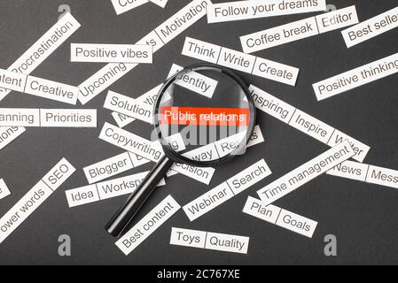 Lupenglas über der roten Aufschrift Public Relations aus Papier geschnitten. Umgeben von anderen Inschriften auf dunklem Hintergrund. Word Cloud-Konzept. Stockfoto
