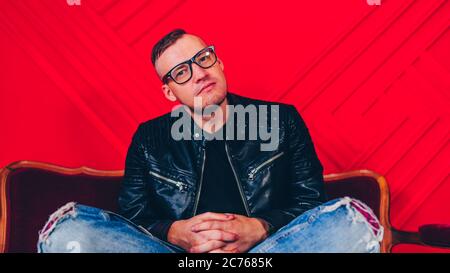Schöner junger Mann mit Brille auf kleinem Sofa. Erwachsene charmante männliche posiert, sitzt auf Sessel gegen rote Wand. Stockfoto