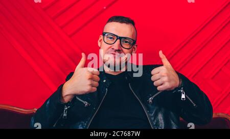 Schöner junger Mann mit Brille zeigt Daumen nach oben auf Hintergrund rote Wand. Erwachsene junge Mann auf dem Sofa sitzend Geste der Zustimmung. Das Konzept von Stockfoto