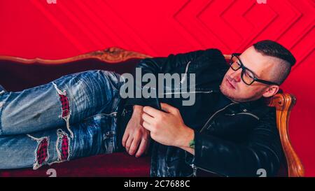 Hübscher junger Mann mit Brille, die im Handy auf einem kleinen Sofa an der roten Wand ruht. Erwachsene charmante männliche Flipping durch verschiedene Nachrichten in mobile Stockfoto