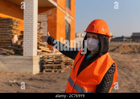 Bauarbeiterin in Overalls und in medizinischer Maske, die auf das Haus im Bau zeigt. Stockfoto