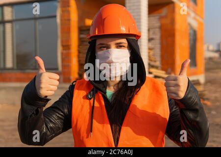 Bauarbeiterin in Overalls und in medizinischer Maske zeigt Daumen nach oben auf dem Hintergrund des Hauses im Bau. Junge Frau in Hut und Stockfoto