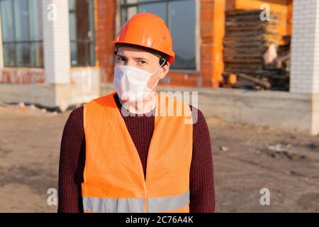 Porträt des männlichen Bauarbeiters in medizinischer Maske und Overalls auf dem Hintergrund des Hauses im Bau. Stockfoto