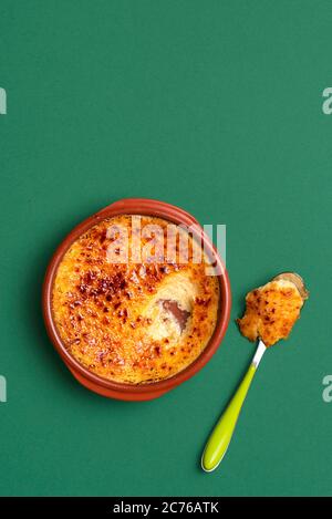 Französisch Crème brûlée Dessert in einem Tonteller mit Löffel, auf grünem Hintergrund. Draufsicht auf gebrannte Sahne mit Karamellkruste. Pudding mit verbranntem Zucker. Stockfoto