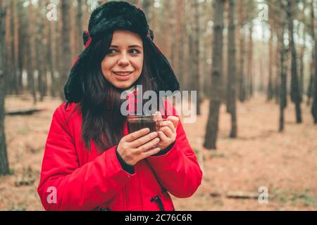 Ein Porträt der hübschen Frau mit einer Tasse Tee im Wald. Die Frau in einer Jacke und einem Hut mit Ohrenklappen trinkt bei kaltem Wetter einen Tee. Stockfoto