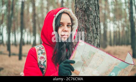 Eine Reisende Frau mit Karte im Wald. Ein Porträt der hübschen Frau mit Rucksack, die bei kaltem Wetter in der Nähe eines Baumes steht. Stockfoto