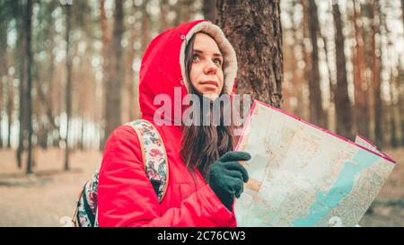 Eine Reisende Frau mit Karte im Wald. Ein Porträt der hübschen Frau mit Rucksack, die bei kaltem Wetter in der Nähe eines Baumes steht. Stockfoto