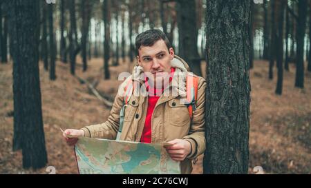 Ein Reisender Mann mit Karte im Wald. Ein Porträt des gutaussehenden Mannes mit Rucksack, der bei kaltem Wetter in der Nähe eines Baumes steht. Stockfoto