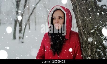 Porträt einer jungen Frau in roter Jacke und Kapuze in der Wintersaison. Schöne Dame steht neben Baum und schaut weg. Flauschiger Schnee umhüllt alles Stockfoto